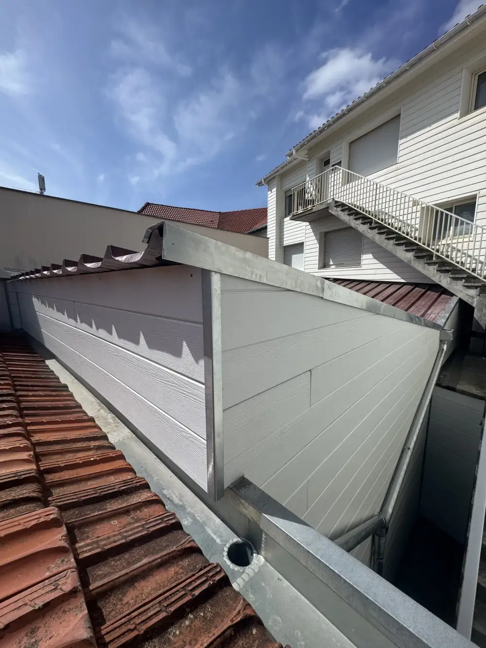 Bardage clair à lames horizontales encadré de profilés métalliques, avec chéneau et descente d’eau près d’un toit de tuiles