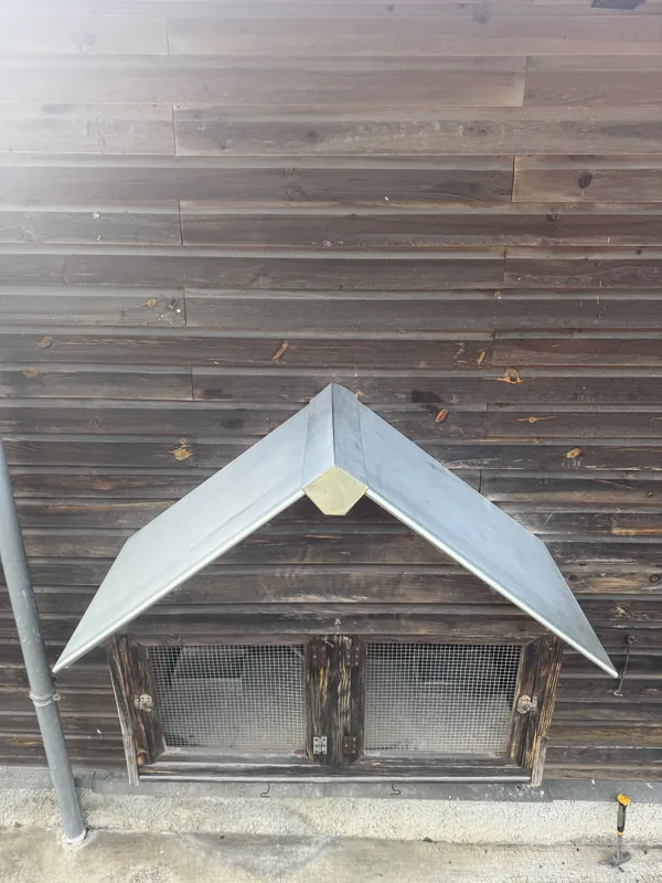 Petit auvent à deux pans en métal gris au-dessus d’une ouverture grillagée, sur un mur en planches de bois