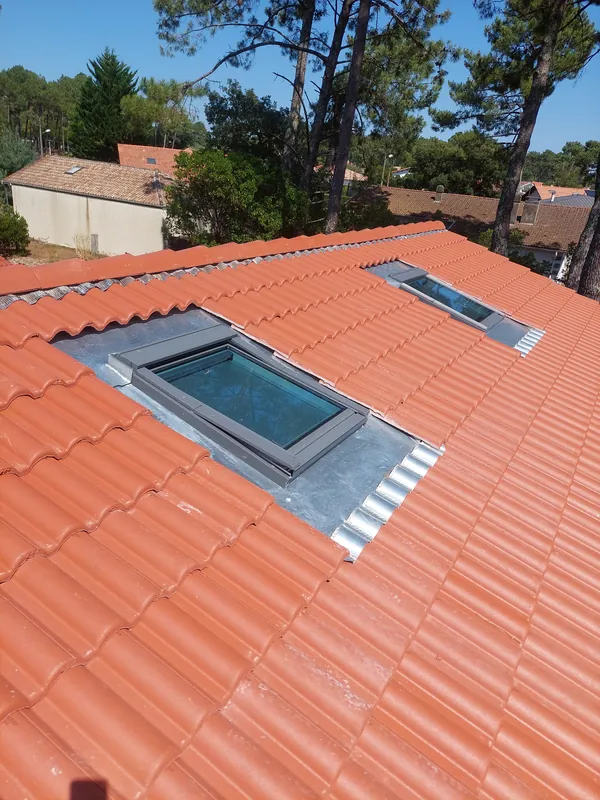 Deux fenêtres de toit encadrées de bandes de métal gris sur un toit de tuiles rouge-orangé