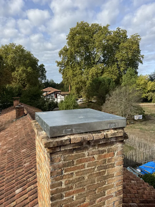 Souche de cheminée en briques coiffée d’une plaque de métal gris à rebords, sur un toit de tuiles anciennes